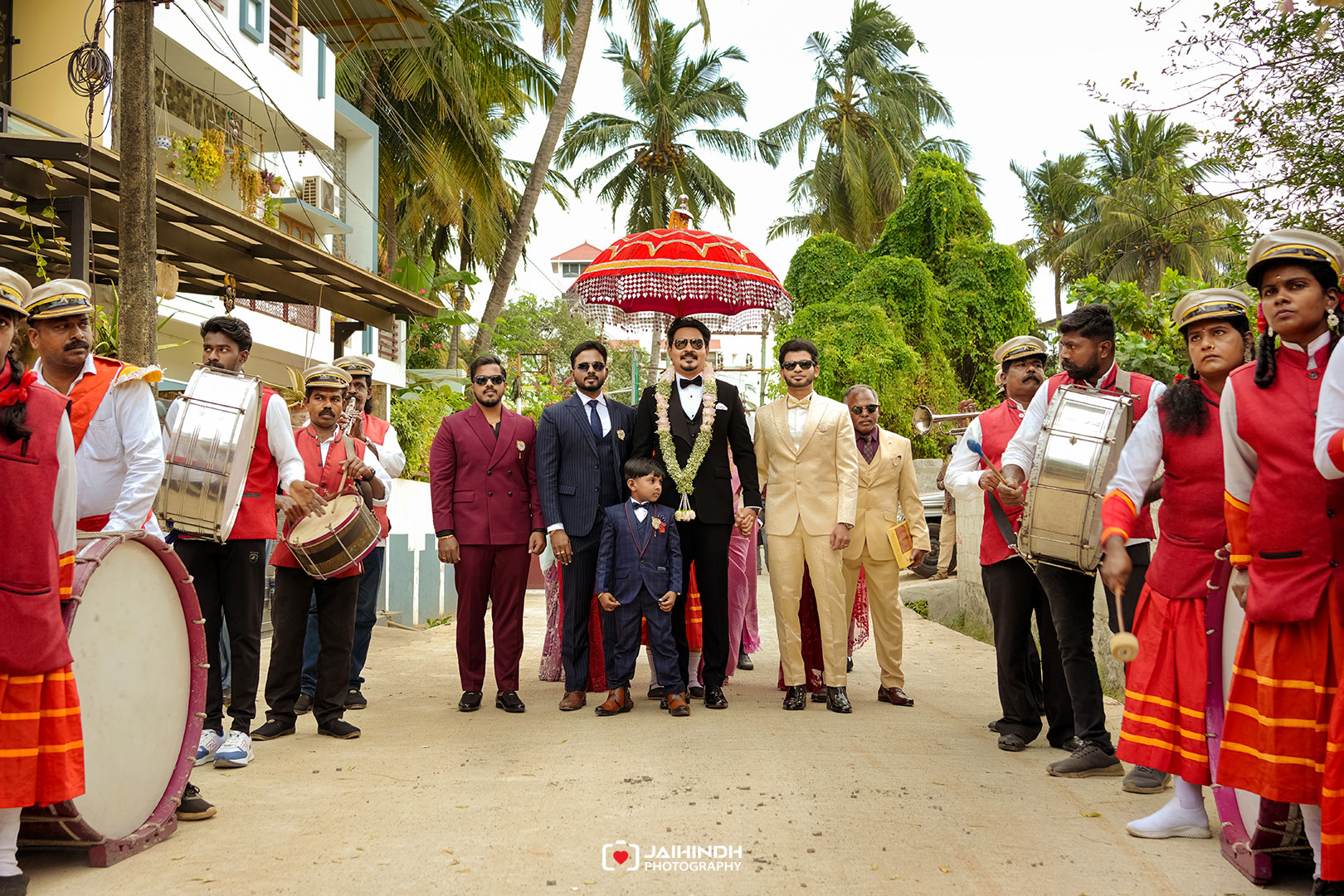 Groom in a black suit with garland under a red decorative umbrella, flanked by family and musicians in a street wedding procession.