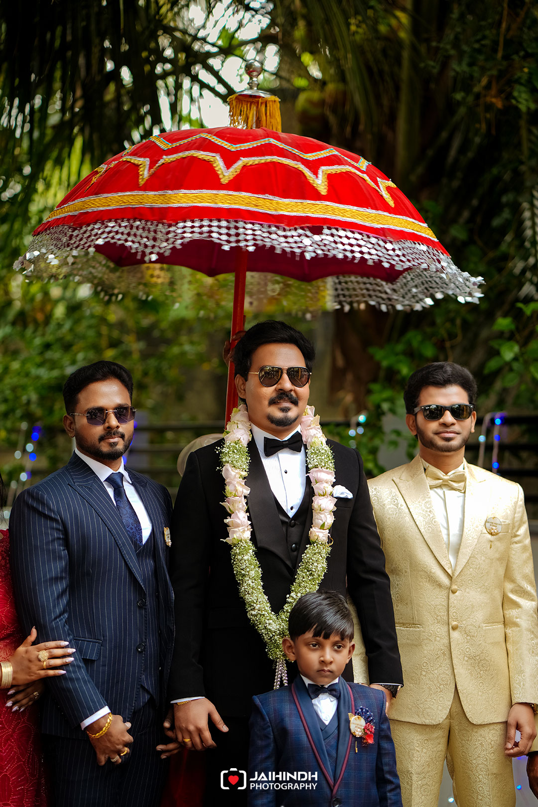 Groom in a black tuxedo under a red decorative umbrella, wearing a floral garland, flanked by suited men and a child in front.