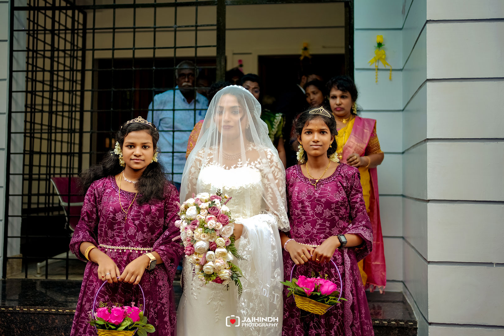 Bride in a white wedding gown with veil holding a bouquet, flanked by two young bridesmaids in purple dresses with flower baskets.