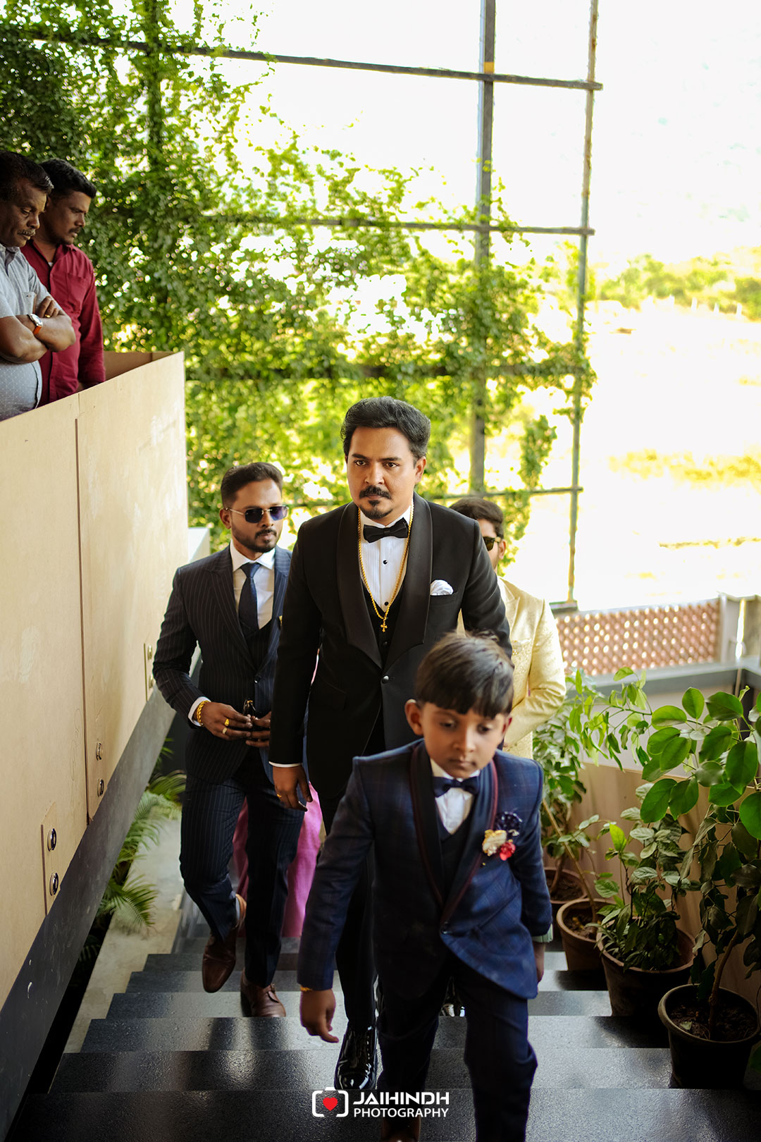 Groom in a black tuxedo leading a line of men in suits down an outdoor staircase, with a young boy in front and greenery in the background.