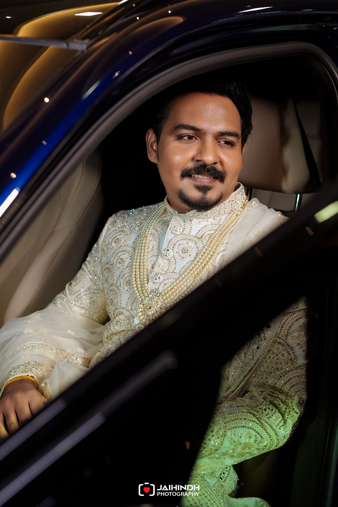 Man in ornate white ceremonial outfit sitting inside a car, looking off to the side, warm jewelry and pearls visible.