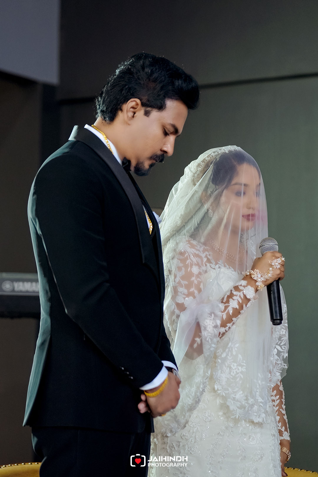 Bride in a lace wedding gown and veil holds a microphone beside a groom in a black suit during a wedding ceremony.