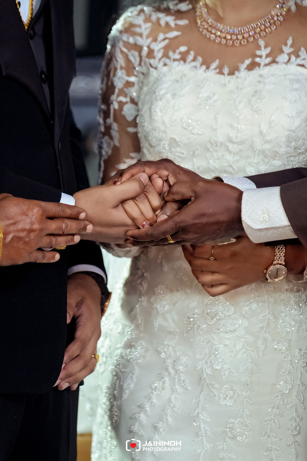 Close-up of a bride and groom joining hands during a wedding, with two other hands guiding and supporting them; lace wedding dress and jewelry in view.