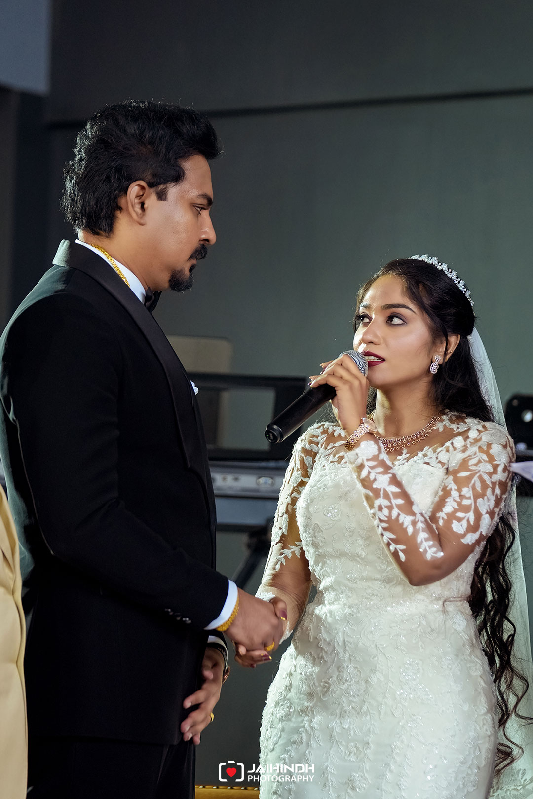 Bride in a white lace wedding gown sings into a microphone while holding the groom's hand; he wears a black suit.