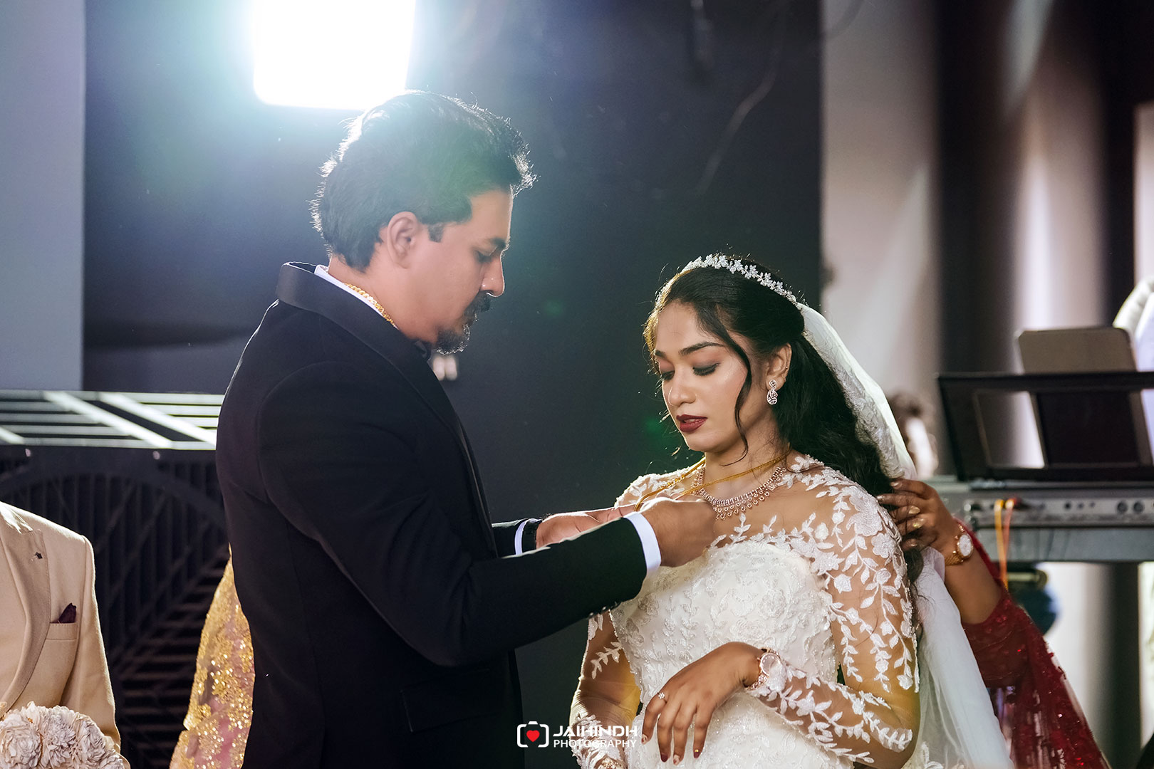 Bride in lace wedding dress and veil, looking down as groom in a dark suit pins her necklace during the ceremony.