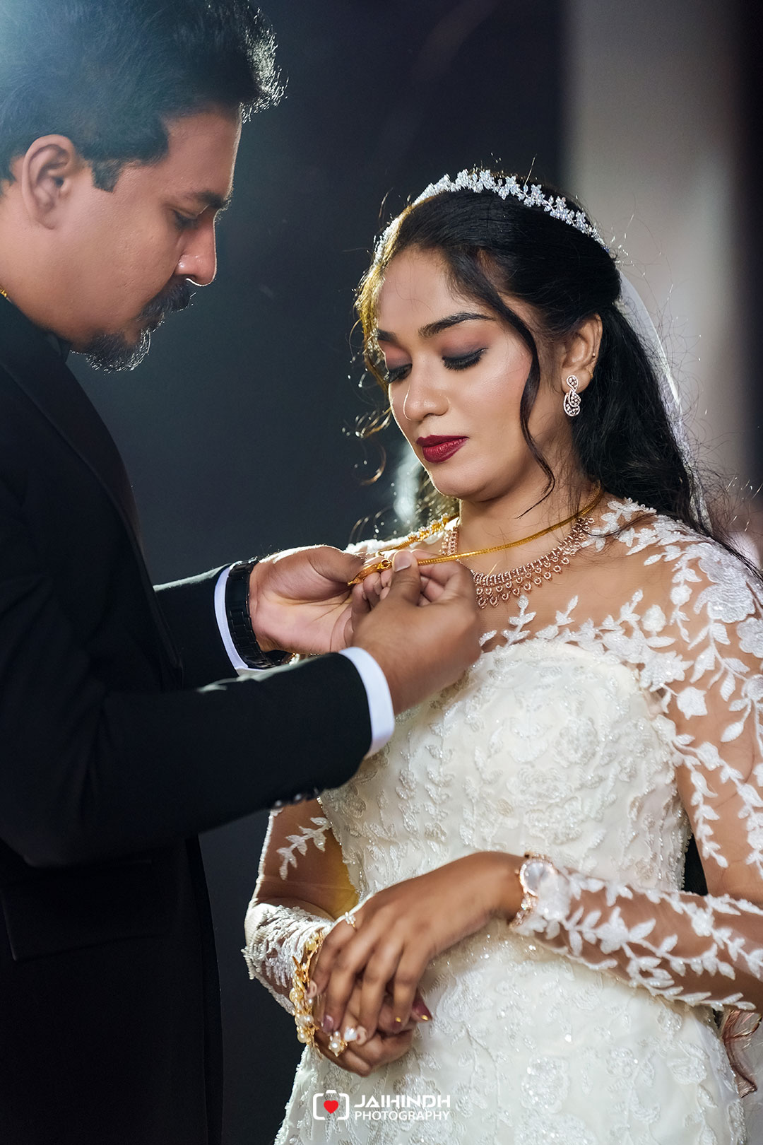 Groom in a black suit fastens a gold necklace on the bride wearing a lace wedding gown and veil.