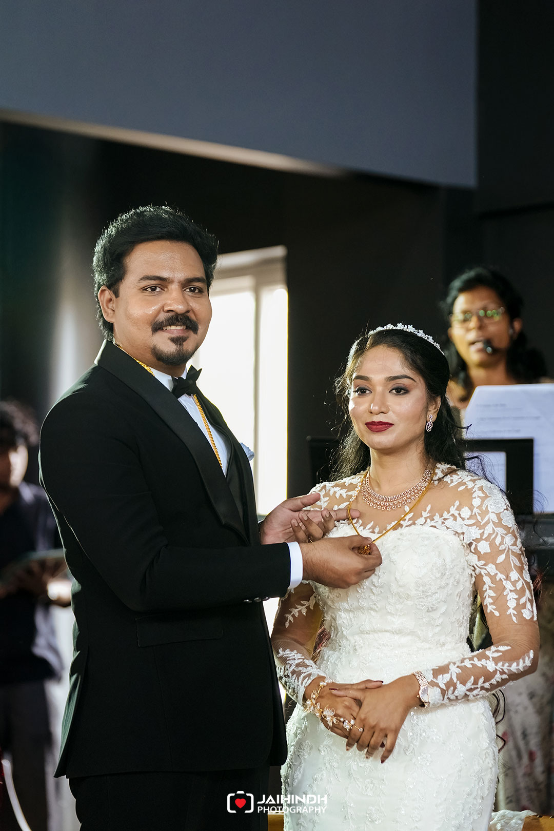 Bride and groom exchange rings during a wedding ceremony; the groom places a ring on the bride's finger as she smiles.