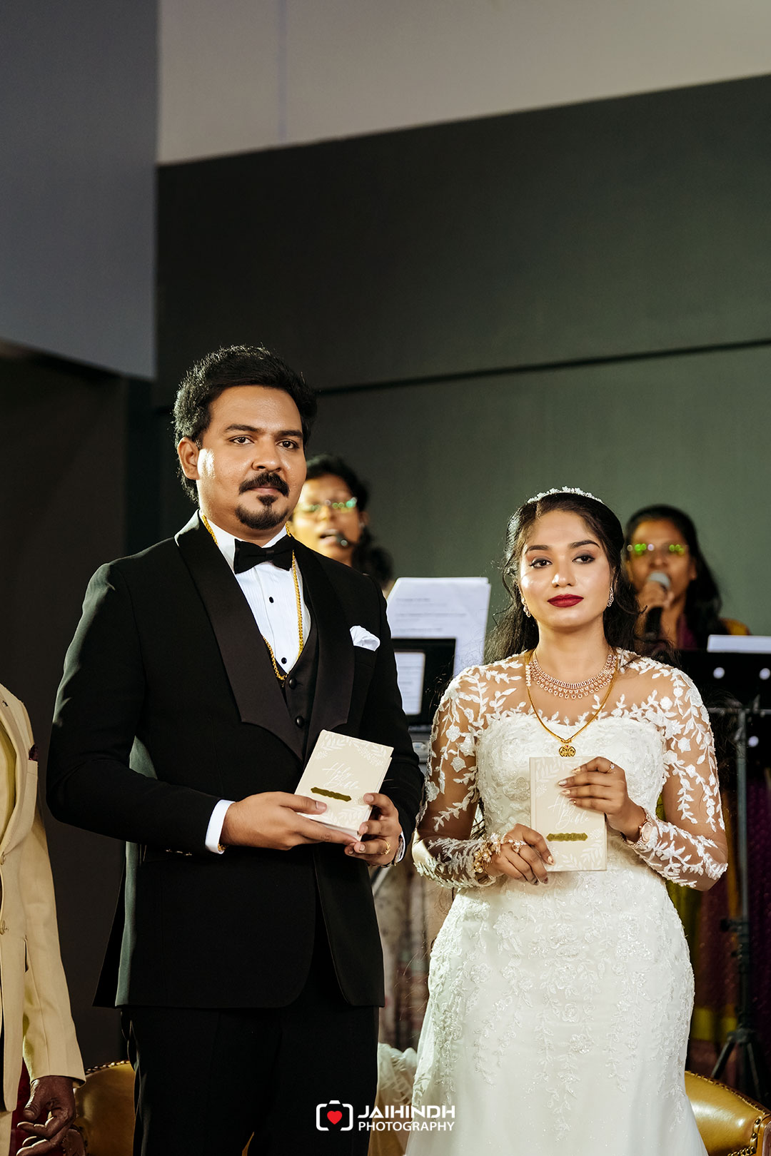 Bride and groom in formal attire holding wedding programs during the ceremony, indoors with a small band in the background.