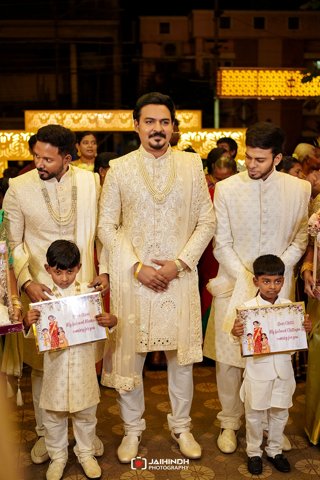 Central groom in an ornate cream sherwani with pearl necklaces stands between two men at a nighttime wedding; two young boys hold invitation cards in the foreground.