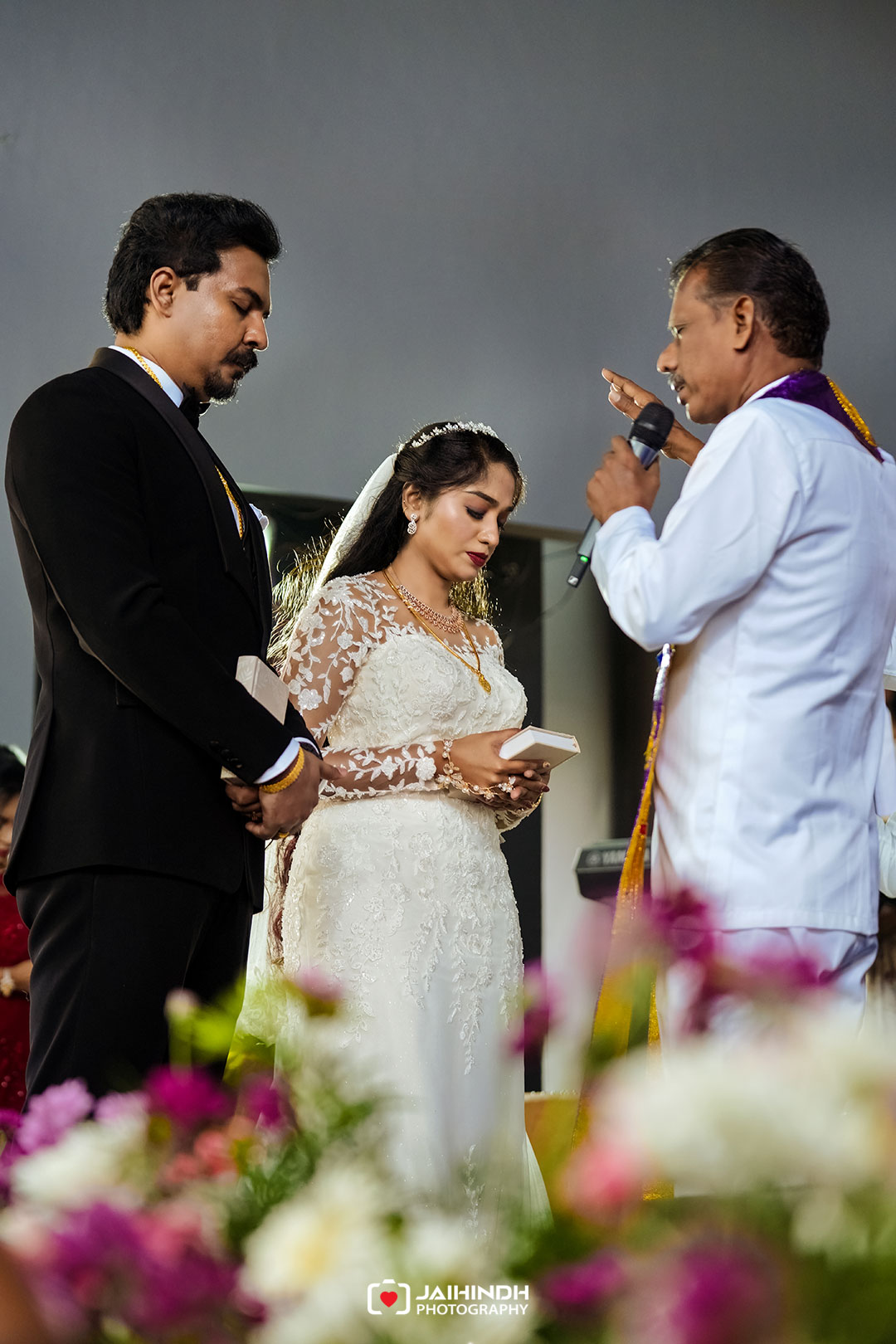 Bride and groom stand with officiant during a wedding ceremony; bride holds a small box while flowers frame the foreground.