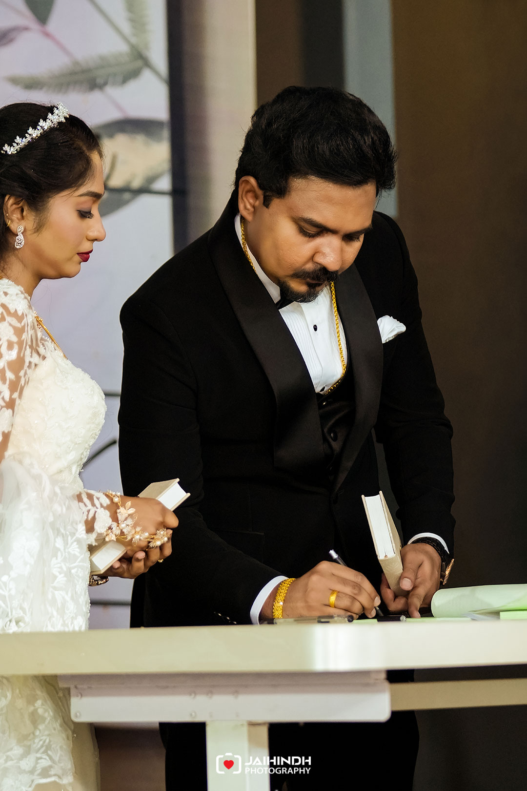 Bride in a lace wedding dress stands beside a man in a black tuxedo as he signs a document at a white table in a formal setting
