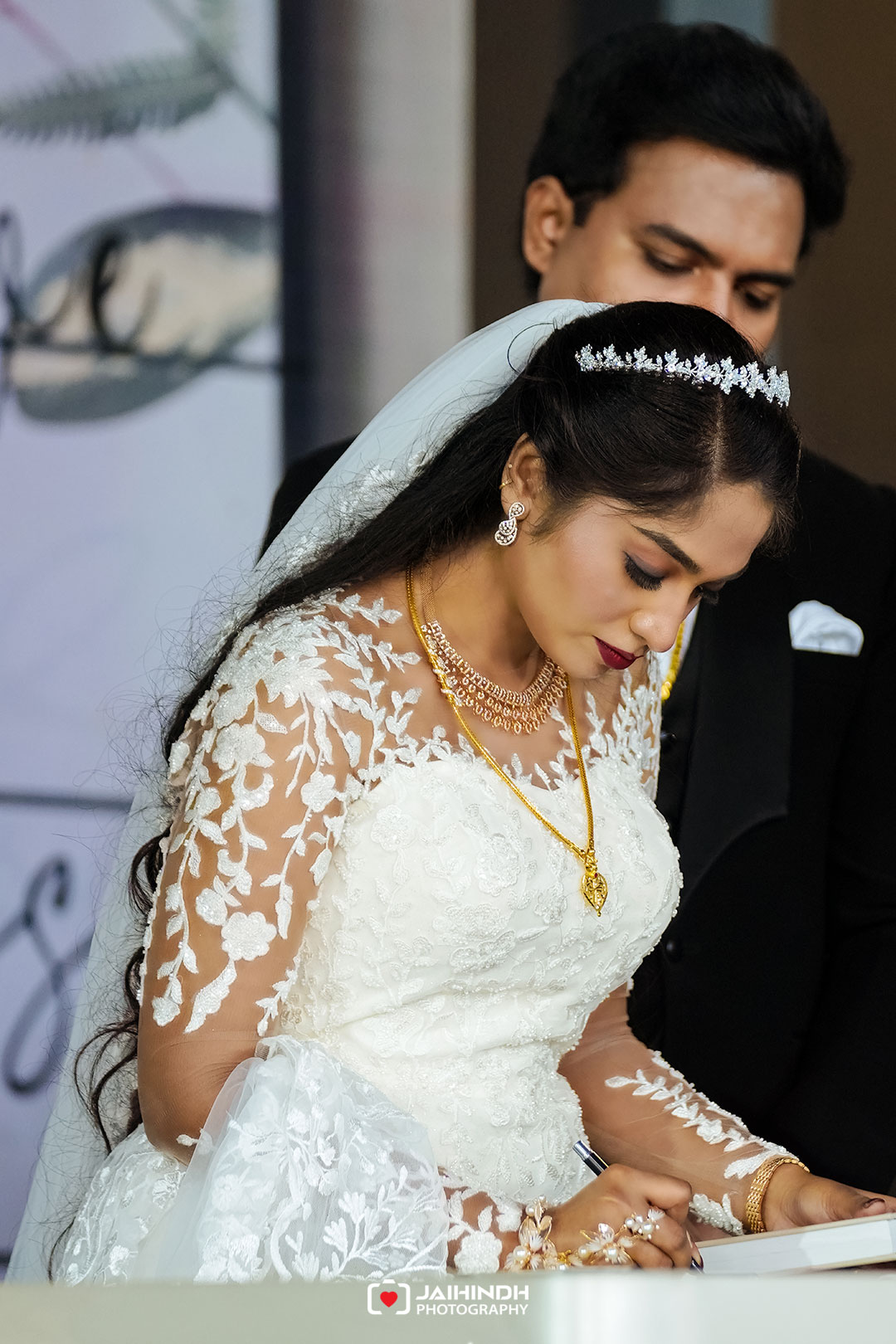 Bride in lace wedding dress signs a guest book, wearing a tiara and veil with gold jewelry, assisted by a man in a black suit behind.