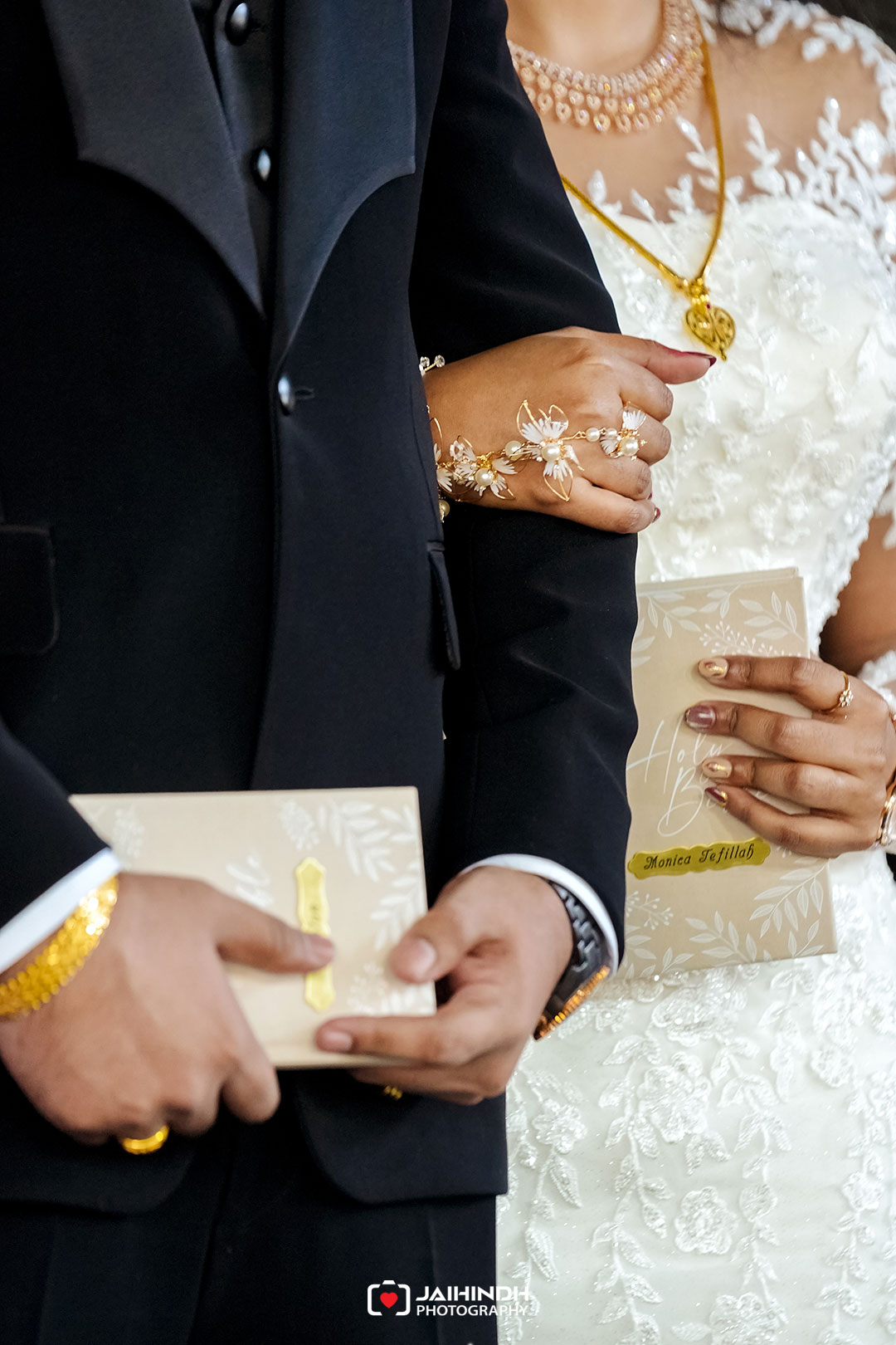 Bride in a lace white wedding dress and gold jewelry holds a beige card while the groom in a black suit stands beside, their hands gently joined.