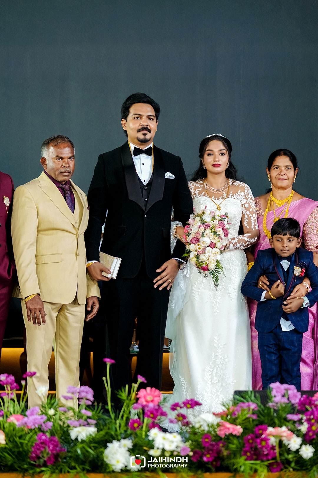 Bride in a white lace gown holds a pink-and-white bouquet, standing beside a man in a black tuxedo; family members in colorful outfits flank them, with flowers in the foreground.