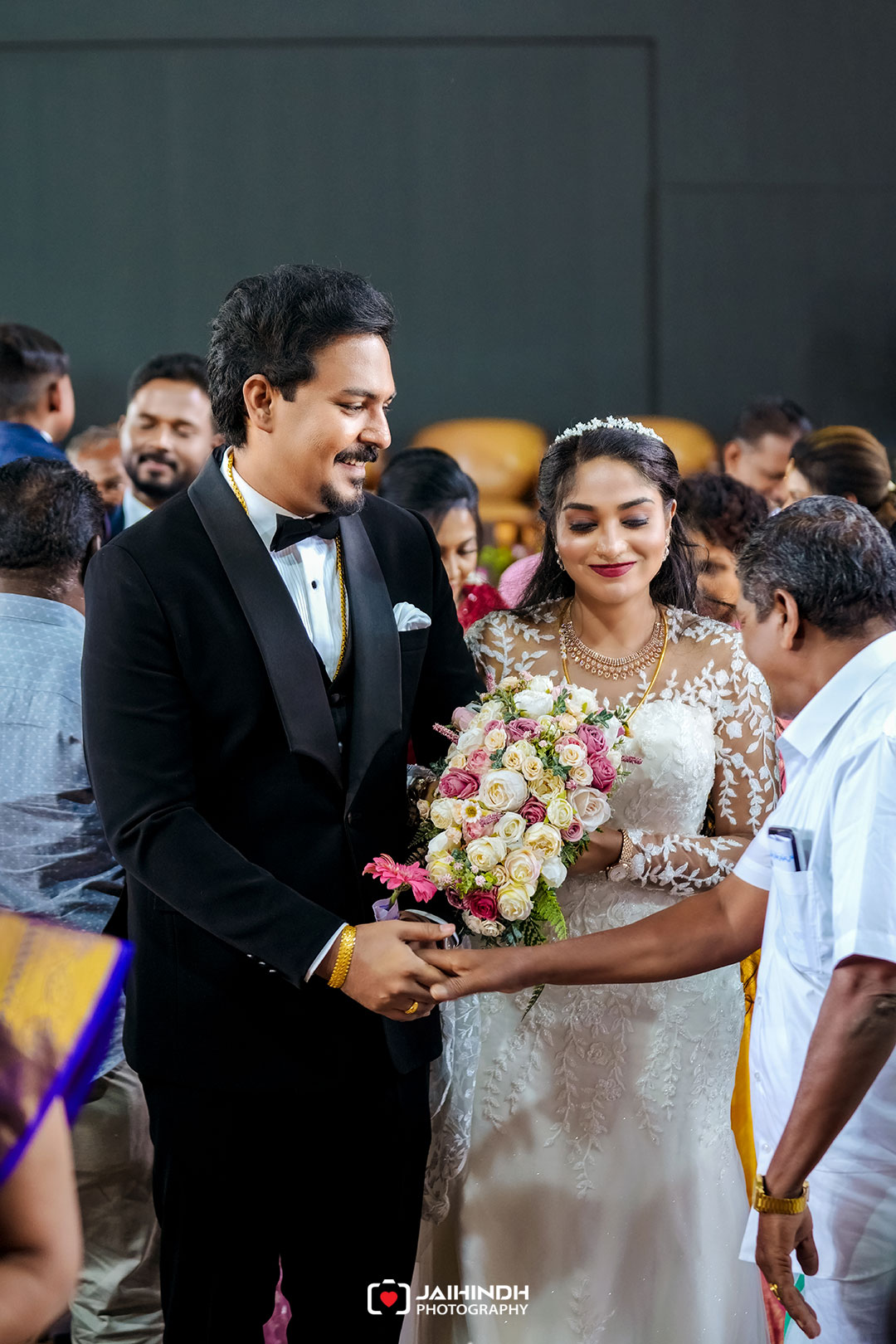 Bride in a lace wedding gown with a tiara and a groom in a black tuxedo smile while greeting a guest; the bride holds a bouquet of white and pink roses.