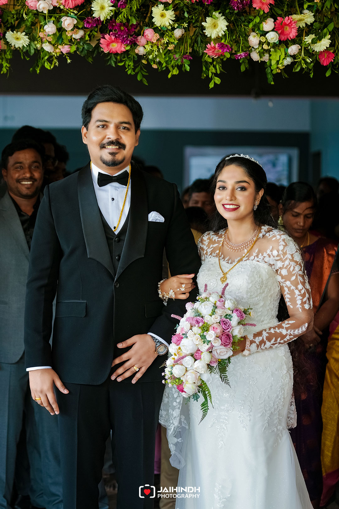 Bride and groom posing arm-in-arm at their wedding under a colorful flower arch, she holds a pink-and-white bouquet.