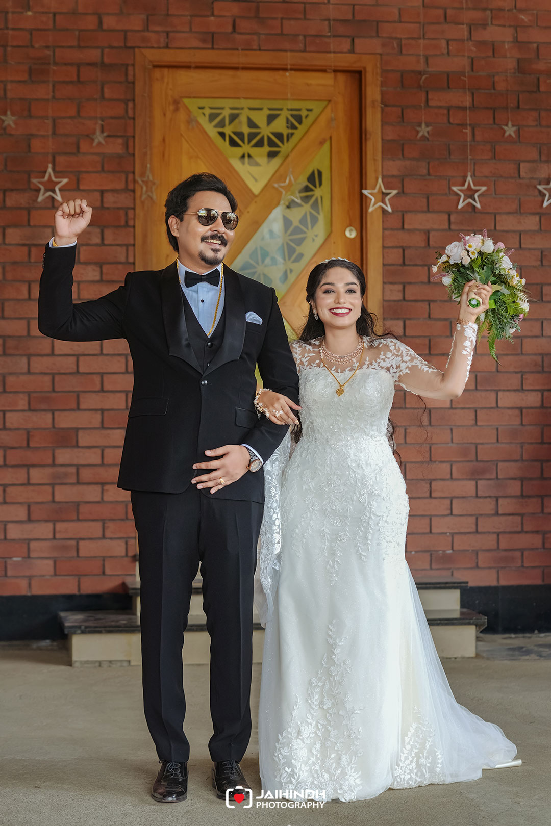 Bride in a lace white wedding gown and groom in a black tuxedo pose arm in arm in front of a brick wall, bride holding a bouquet while smiling at the camera, outdoor setting on steps behind them.