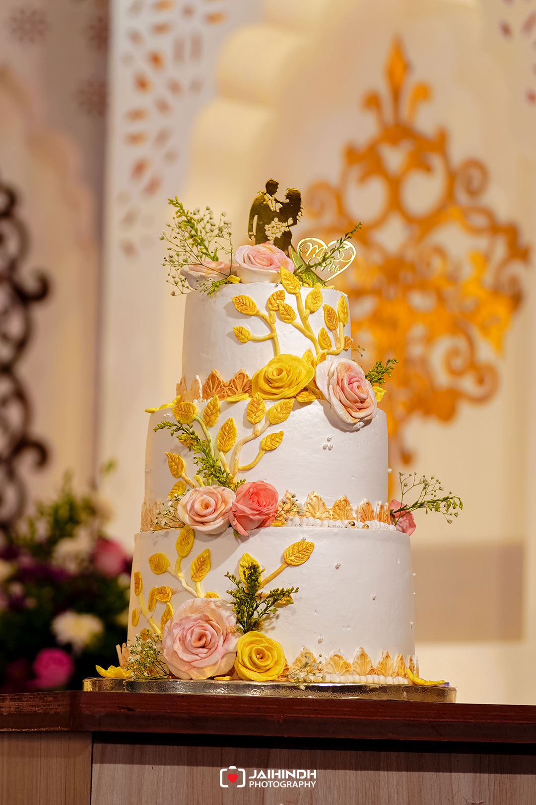 Three-tier wedding cake with gold leaf decorations, pink roses, and delicate greenery, topped by a couple silhouette centerpiece.