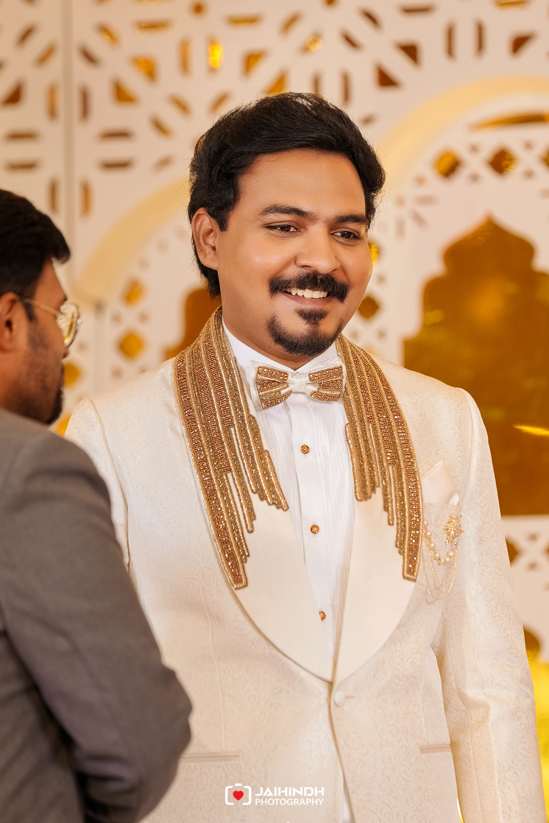 Smiling man in a cream suit with gold embroidered fringe and bow tie at a wedding ceremony.