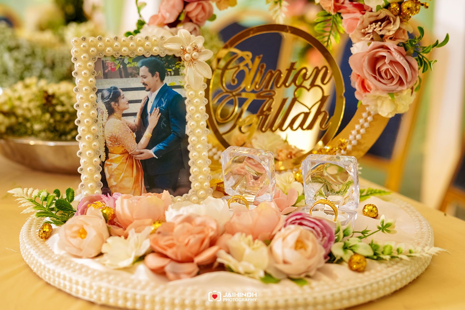 Wedding centerpiece with a pearl-framed photo of the couple, surrounded by pink and white flowers and crystals with rings nearby.