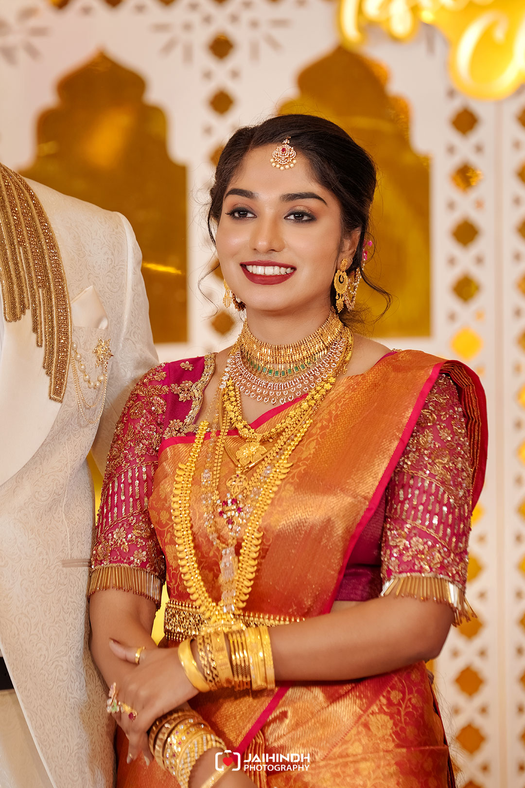 Bride in a red-orange sari with heavy gold jewelry, smiling at the camera during a wedding portrait.
