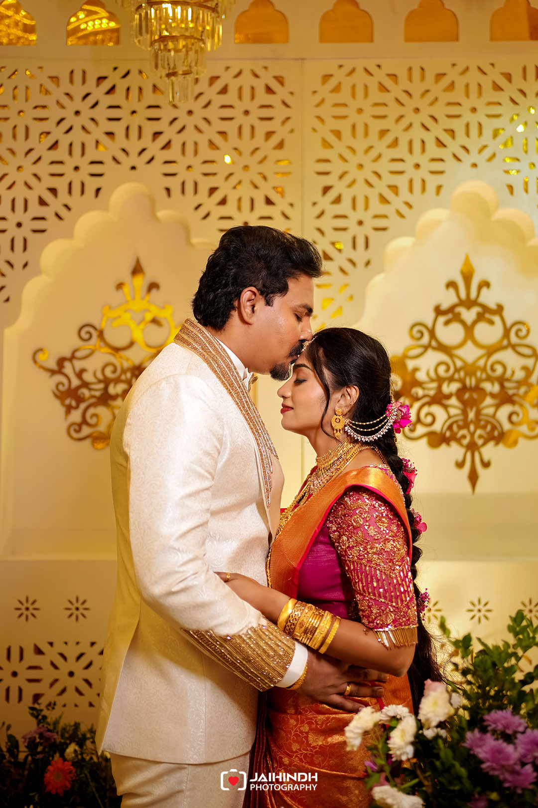 Couple in traditional indian wedding attire share a tender moment, embracing in front of ornate gold decor.
