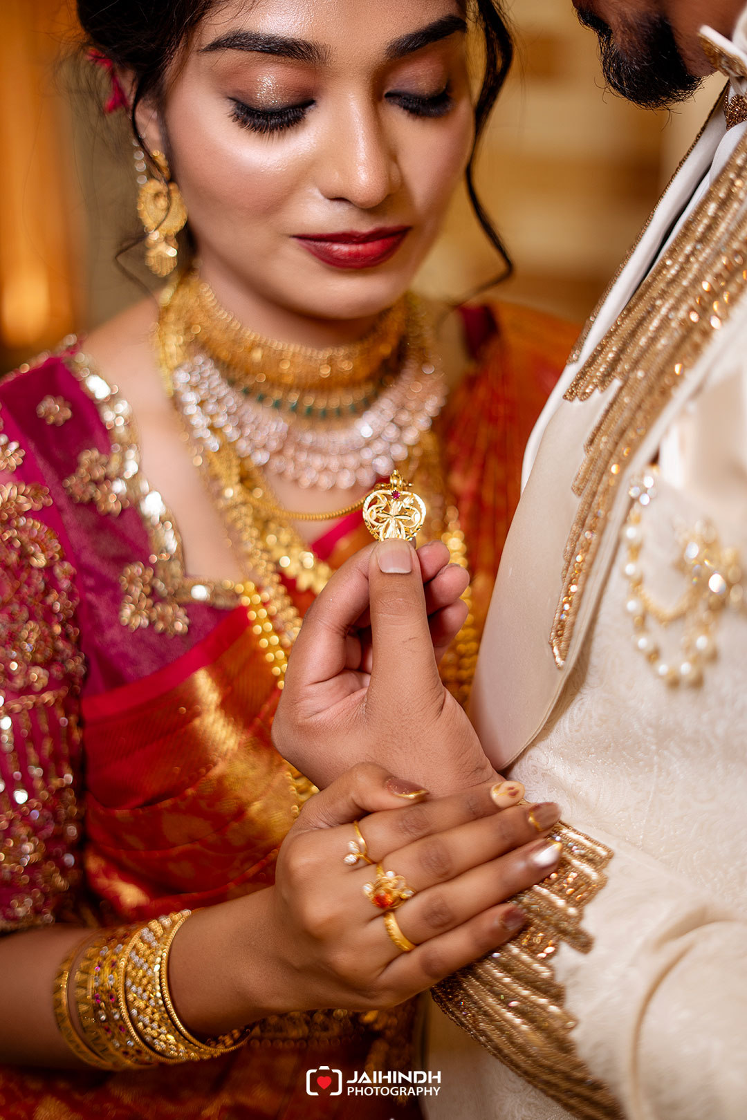 Bride in ornate red and gold attire holds a gold pendant near the groom's cream outfit, showcasing jewelry detail and ceremony mood.