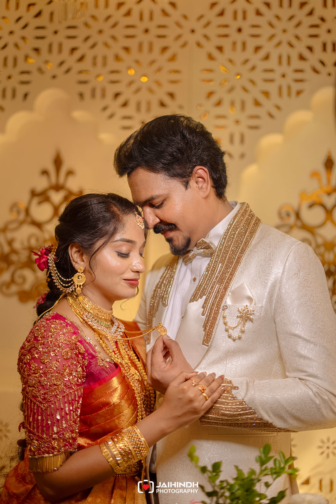 Bride and groom in ornate traditional attire share a tender moment, foreheads touching and hands joined at the wedding.