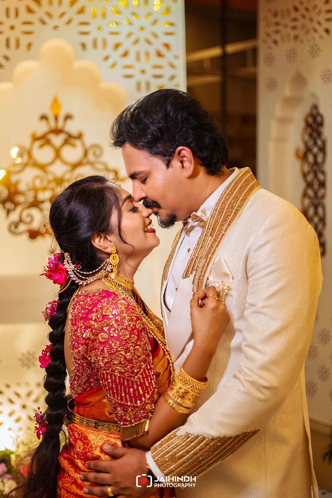 Indian bride and groom share a kiss, dressed in ornate gold and red wedding attire, embracing lovingly.
