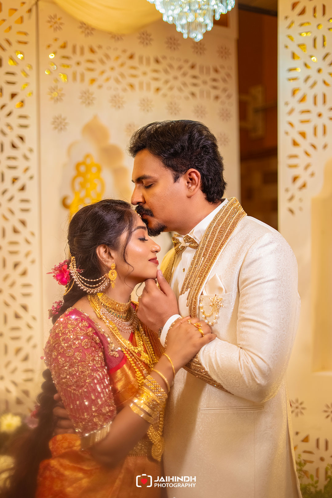 Bride and groom in traditional indian wedding attire embrace; the groom tenderly kisses the bride's forehead in a warmly lit setting.