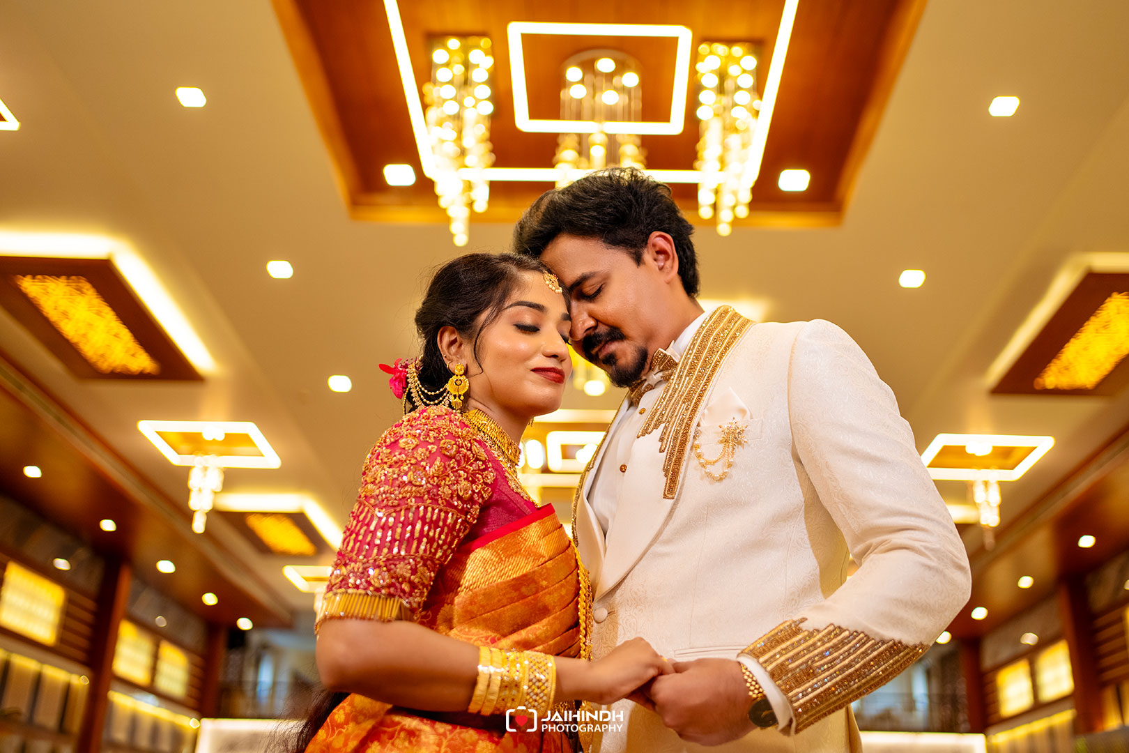 Bride wearing a red-gold sari and ornate jewelry embraces her groom in a cream suit under warm ceiling lights at a wedding venue.