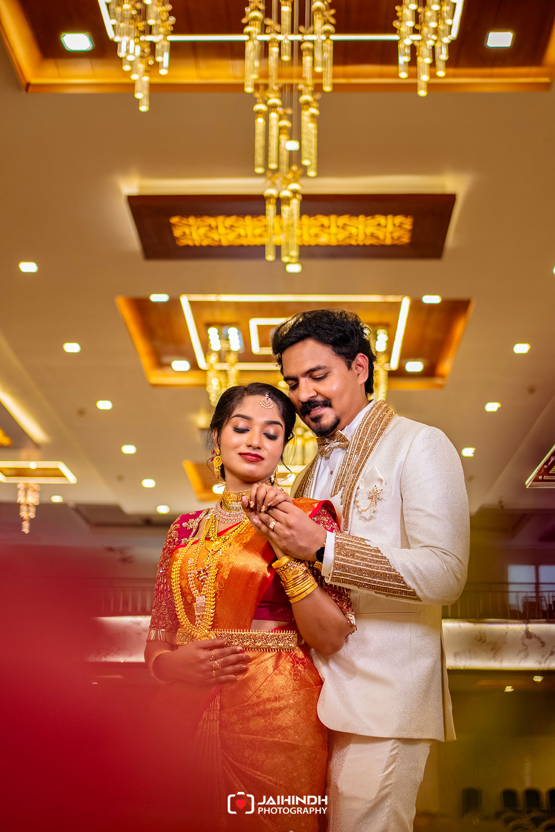 Bride in an orange saree and groom in a cream suit hold hands and smile in a banquet hall with golden chandeliers above.
