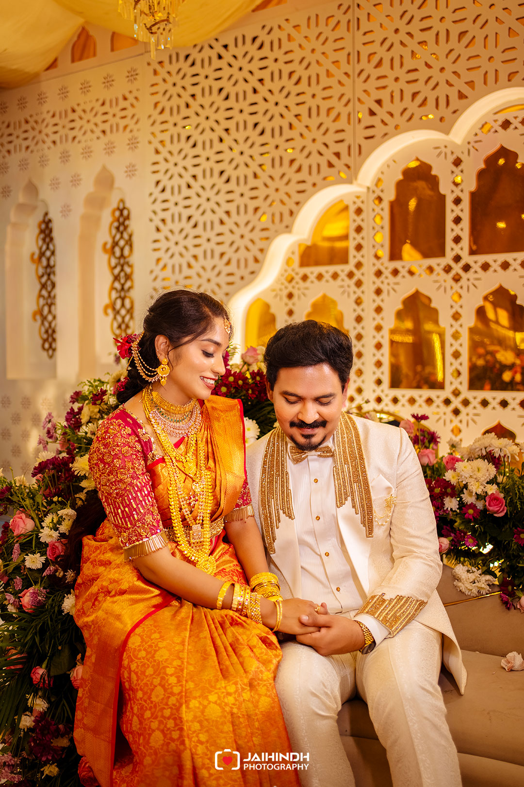 Bride in orange sari sits beside groom in cream suit, holding hands and smiling amid gold decor and flowers.