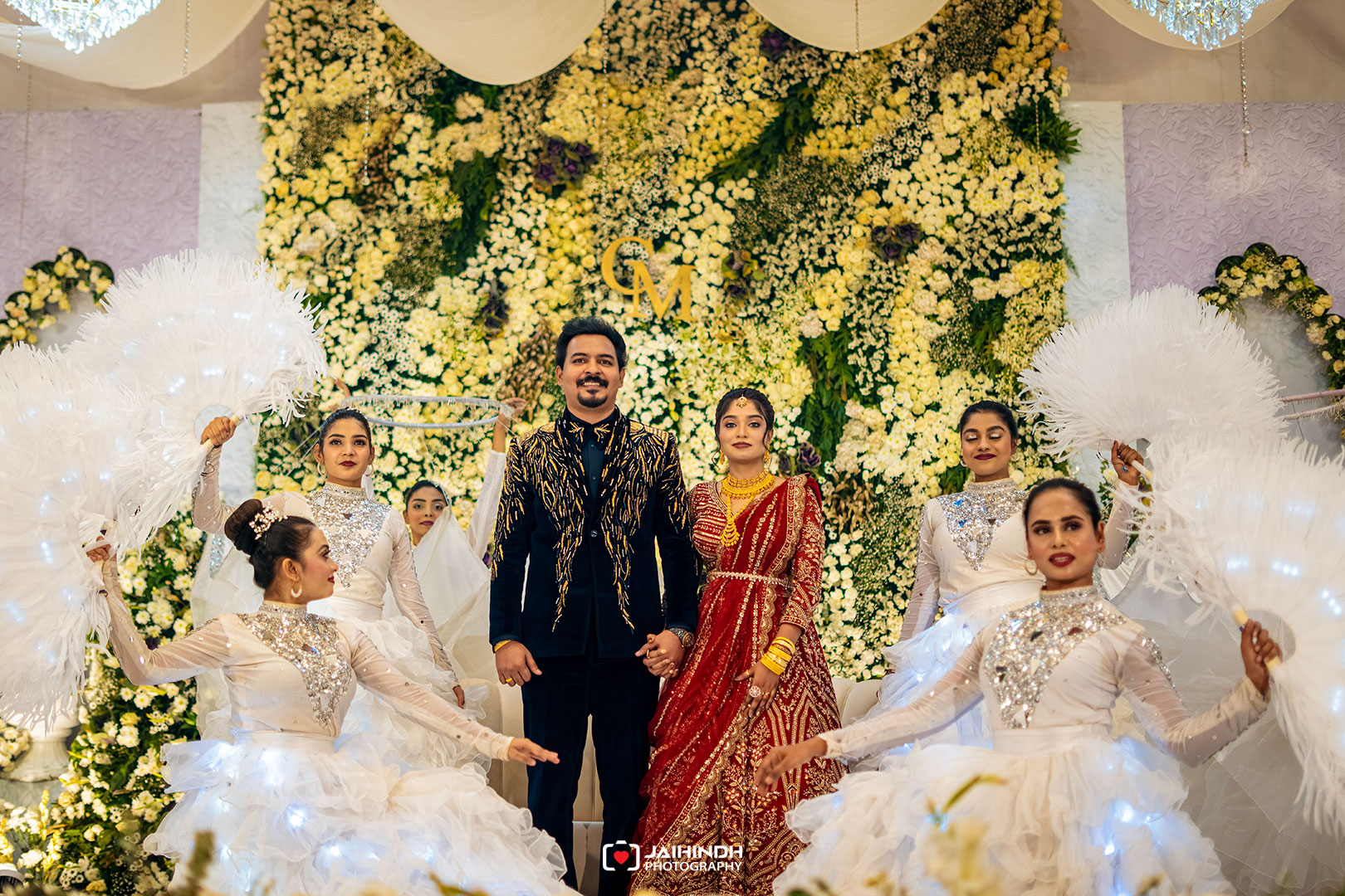 Bride and groom stand hand in hand on a flower-filled stage as dancers in white surround them.