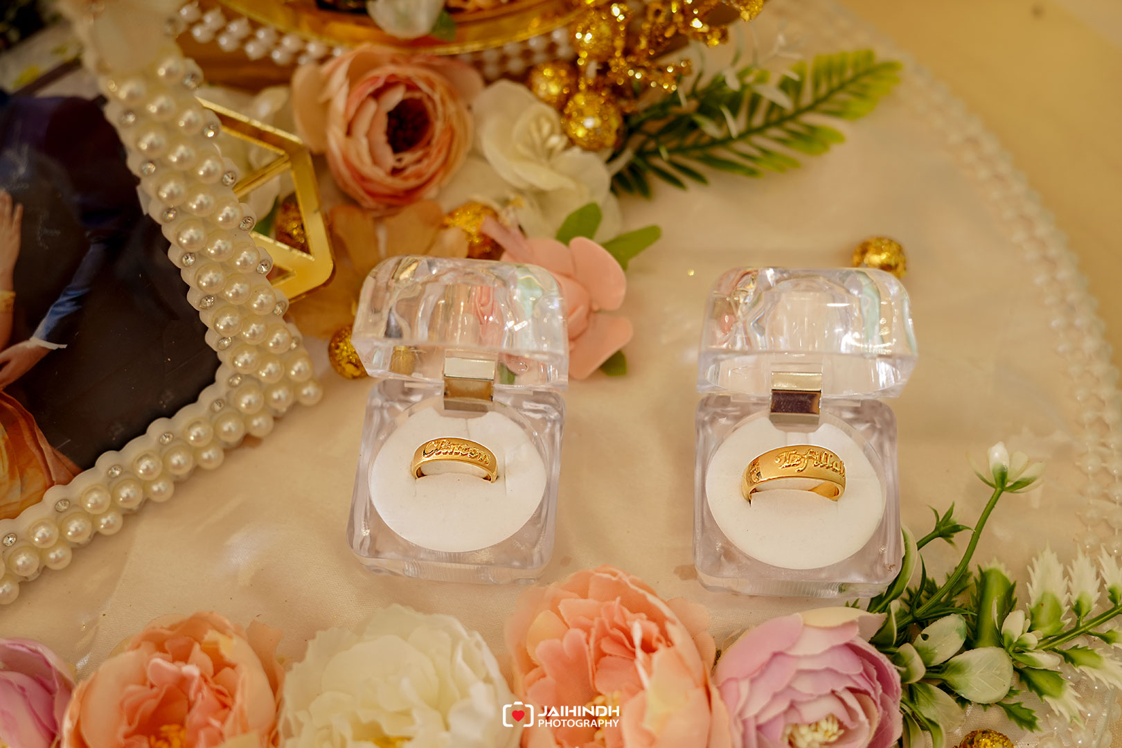 Two gold wedding bands inside clear open ring boxes set on a floral wedding display with pink and white flowers and pearl strands nearby.