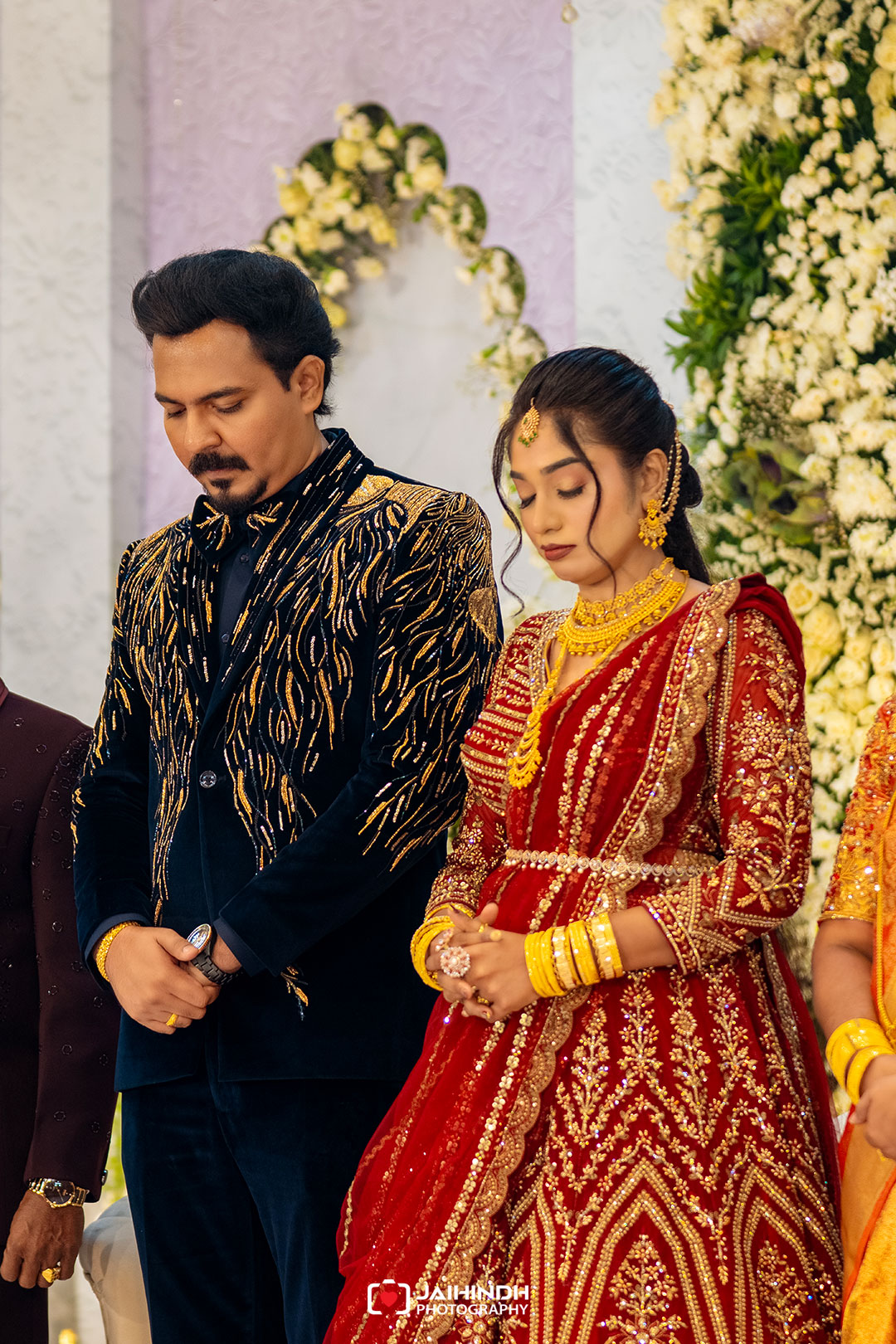 Bride in a red and gold heavily embroidered lehenga with matching jewelry, standing beside a groom in a dark velvet suit at a floral wedding backdrop.