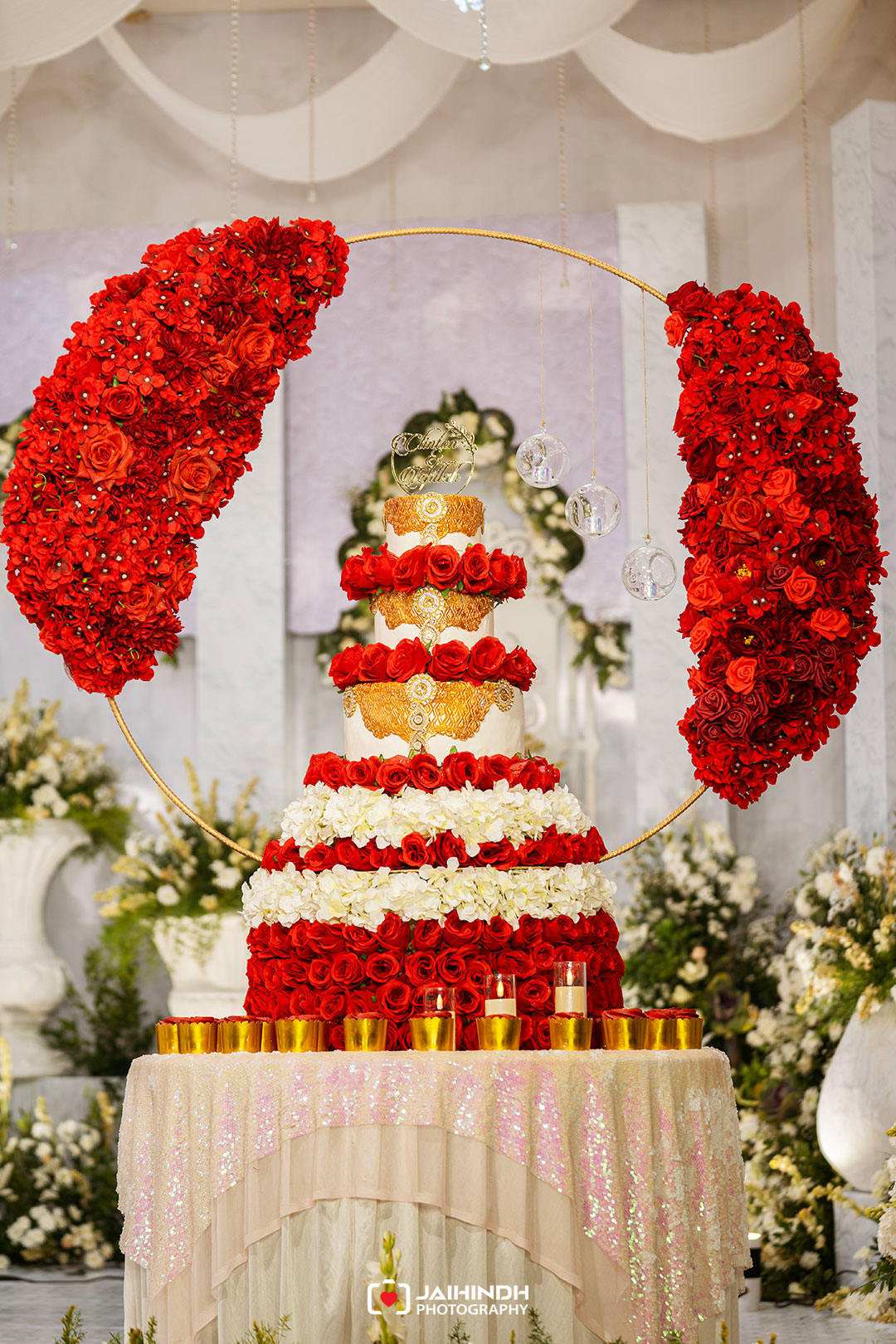 Four-tier wedding cake adorned with red roses and gold lace, beneath a circular arch of red roses and hanging glass orbs for a lavish ceremony display