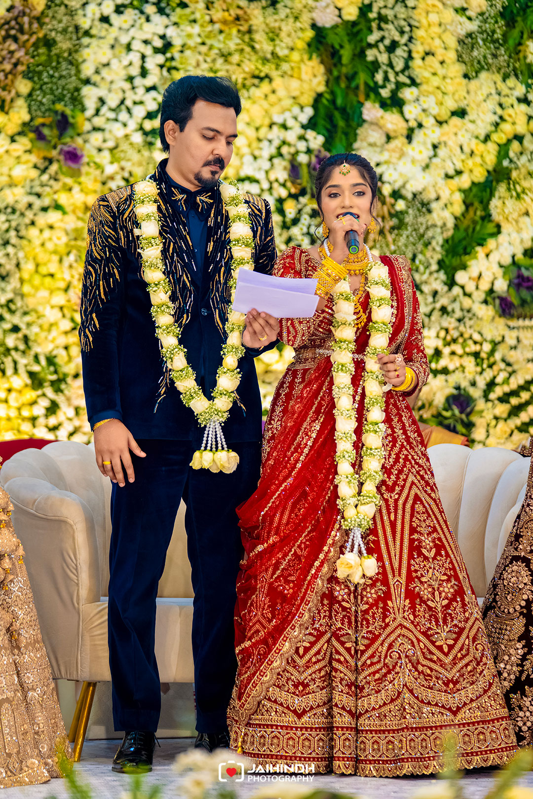 Bride and groom speaking at their wedding, standing with flower garlands in front of a yellow-white floral backdrop.
