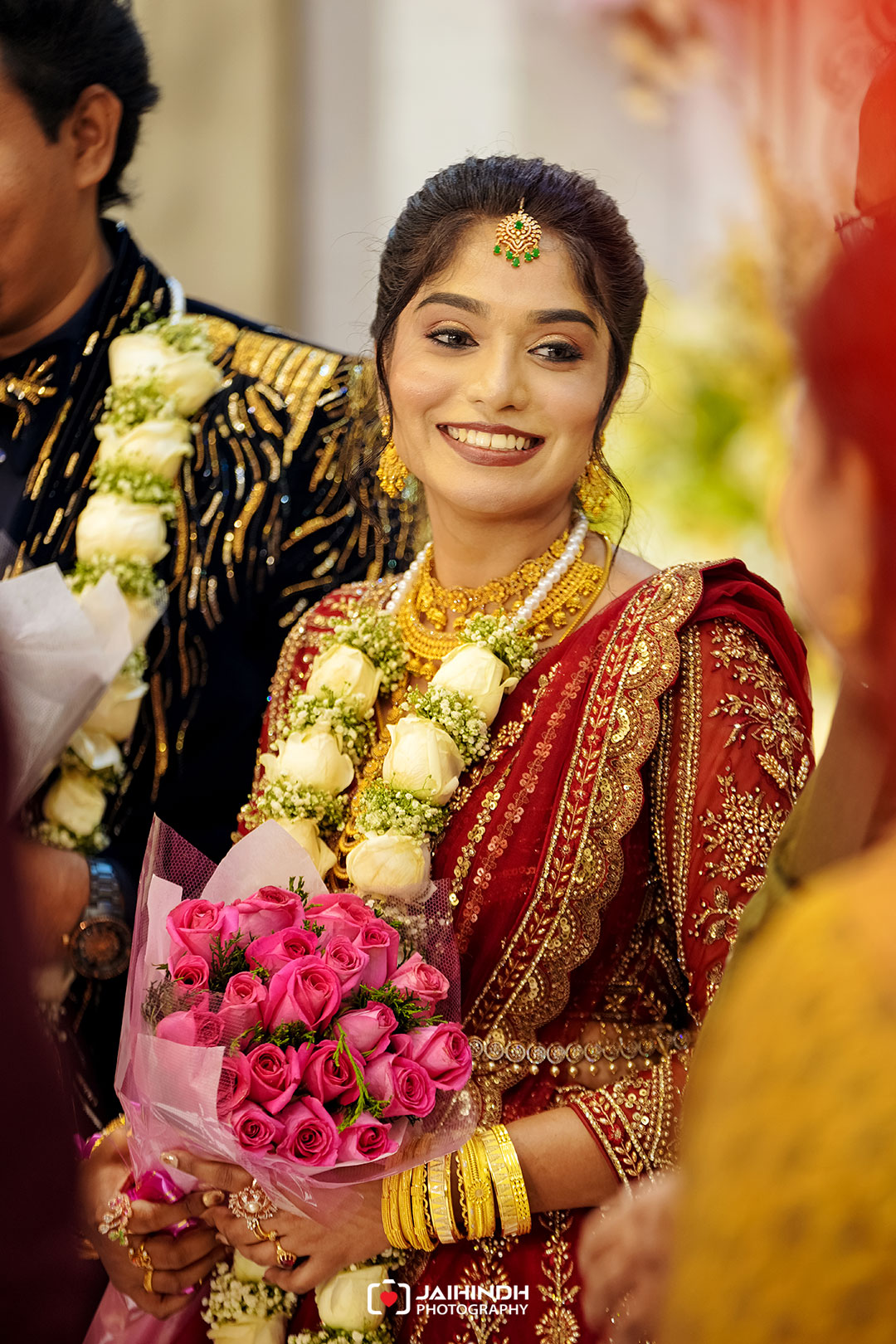 Smiling bride in a red, gold-embroidered saree with heavy gold jewelry, white flower garlands, and a pink rose bouquet.