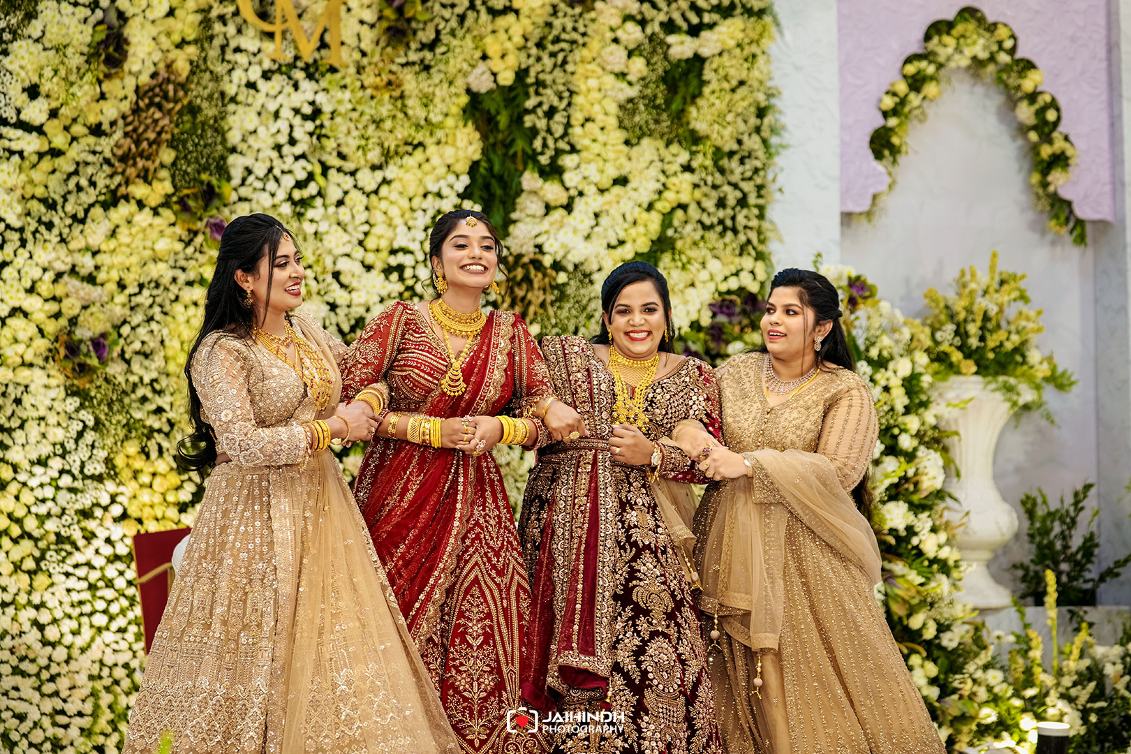 Four women in ornate lehengas posing together at a flower-filled backdrop, smiling and holding hands in a festive celebration.
