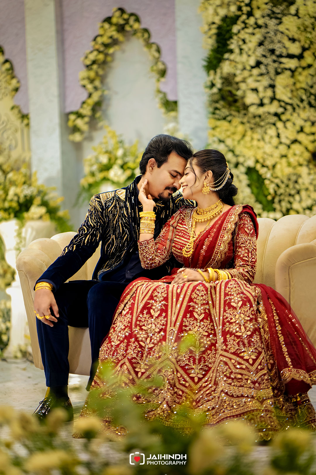 Bride in a red and gold heavily embroidered lehenga sits beside groom in a dark velvet suit, as they lean in for a kiss on a cream sofa with a floral backdrop.