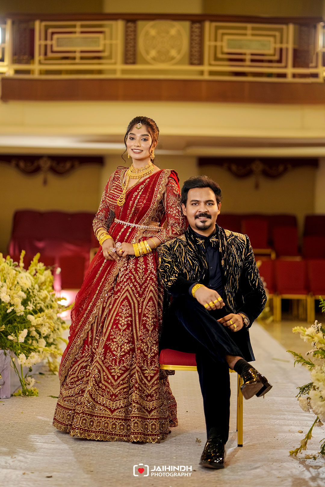 Bride in a red, heavily embroidered lehenga and gold jewelry stands beside a seated groom in a black-gold sherwani at a wedding stage.