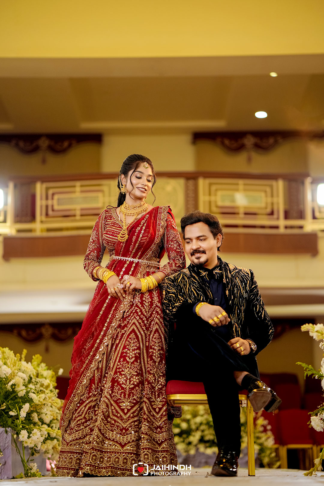 A couple in traditional indian wedding attire posing together on stage—woman in a red embroidered lehenga and gold jewelry, man in a black and gold suit, both smiling.