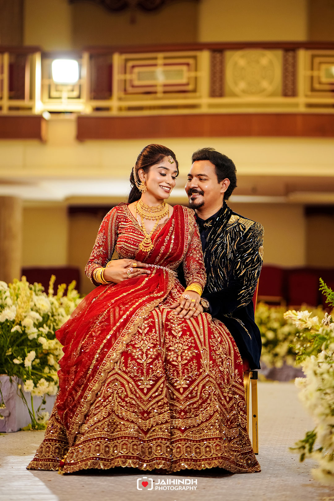 Bride and groom sit close together at a decorated wedding stage, smiling in ornate red bridal attire and a black-gold sherwani.