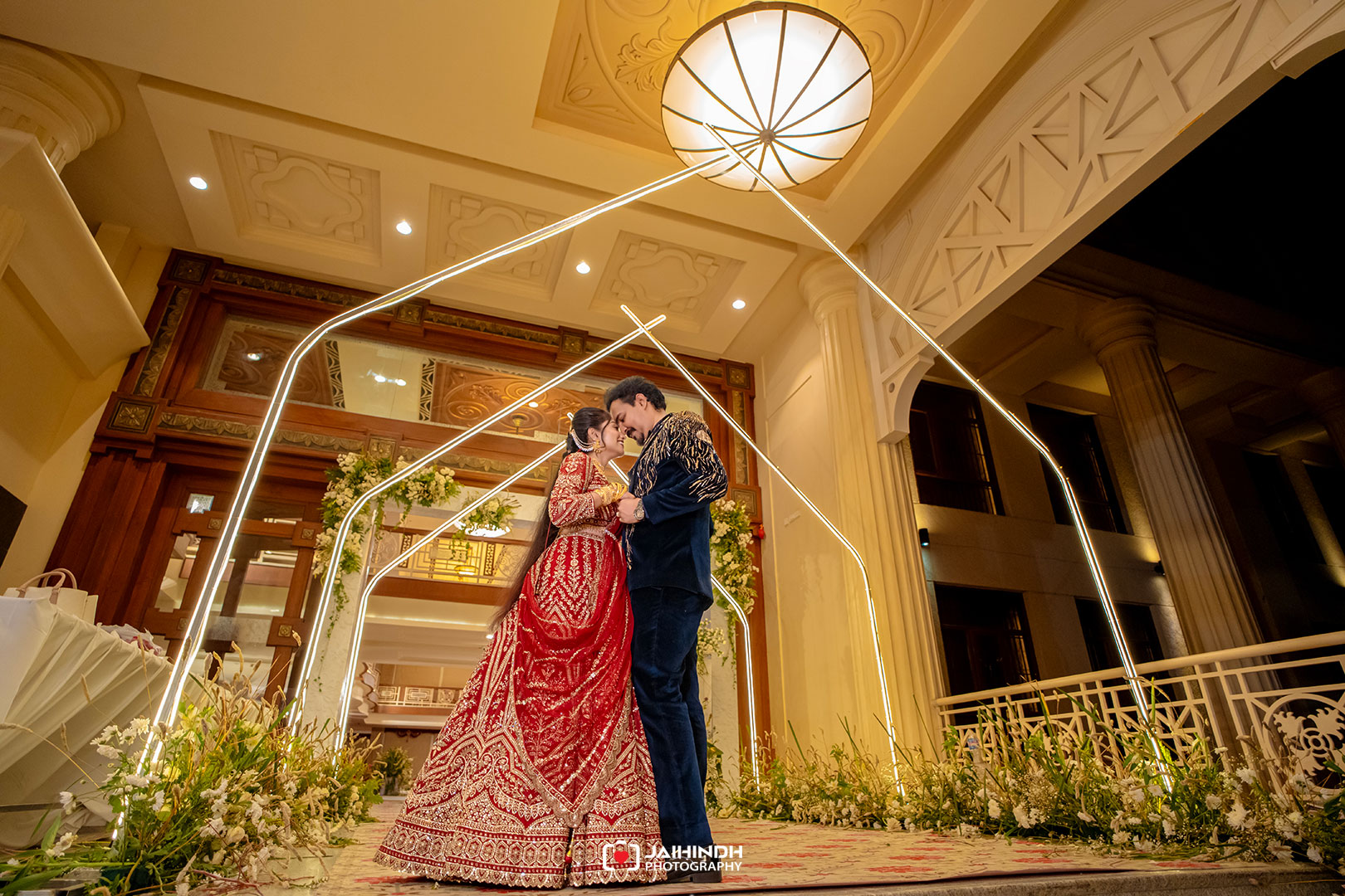 Couple in traditional red wedding attire share a kiss on a lit decorative arch at a luxurious venue.
