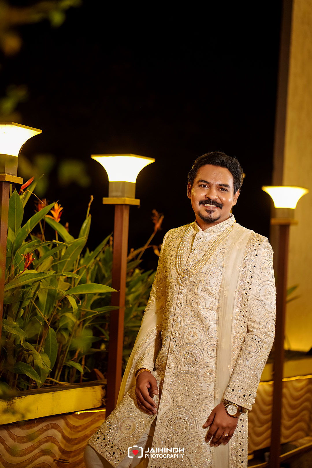 Man wearing an ornate cream sherwani stands smiling outdoors at night beside illuminated lamps on a garden path.