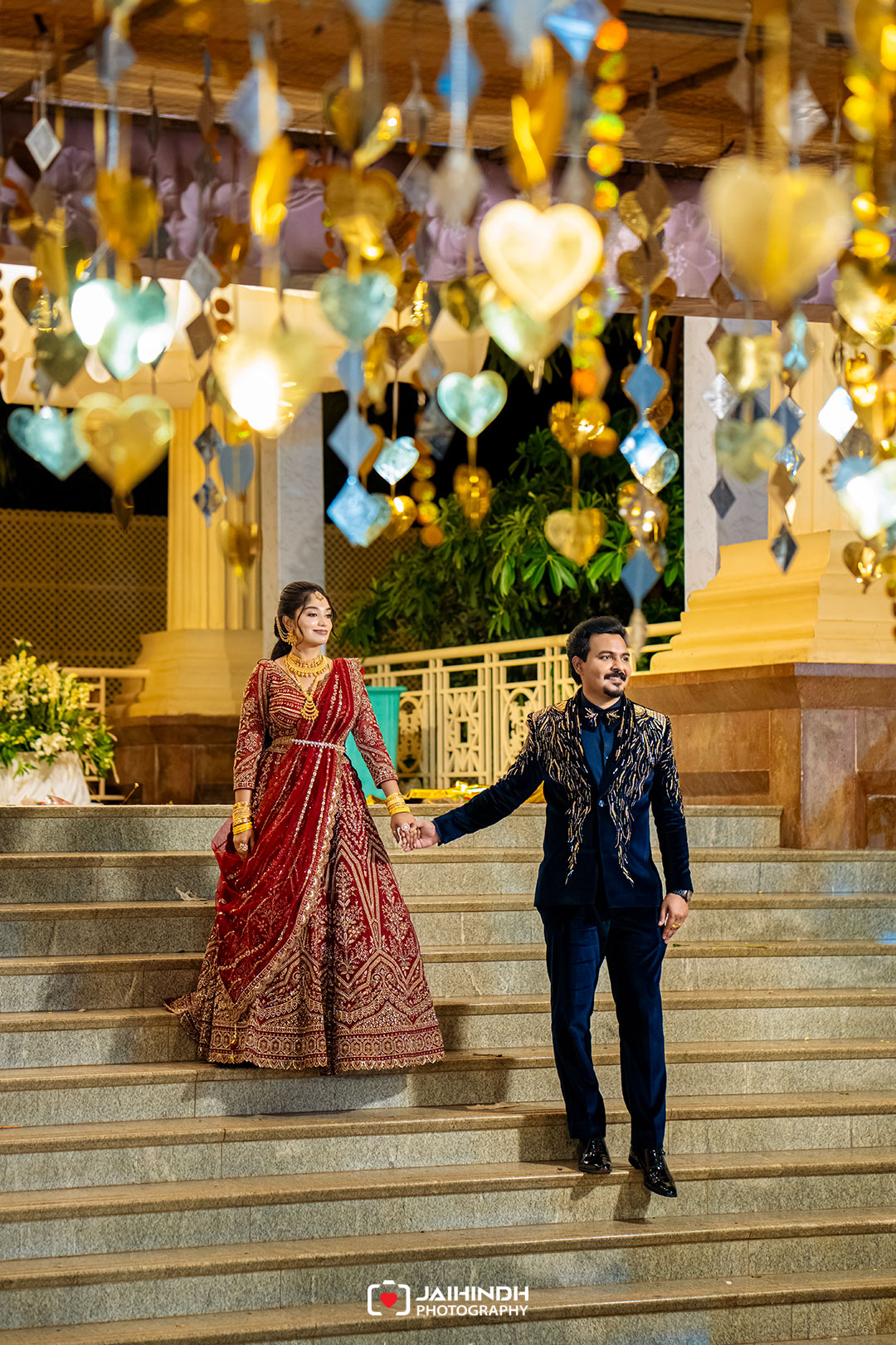 A couple in ornate traditional attire holds hands while standing on stone steps under a ceiling of gold and blue hanging decorations at a festive venue.