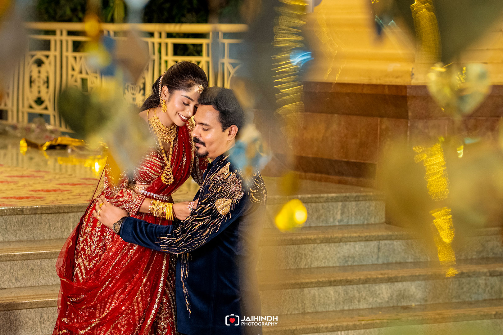 Couple in ornate red and navy outfits embrace on stone steps during a festive indian wedding, smiling warmly at each other.