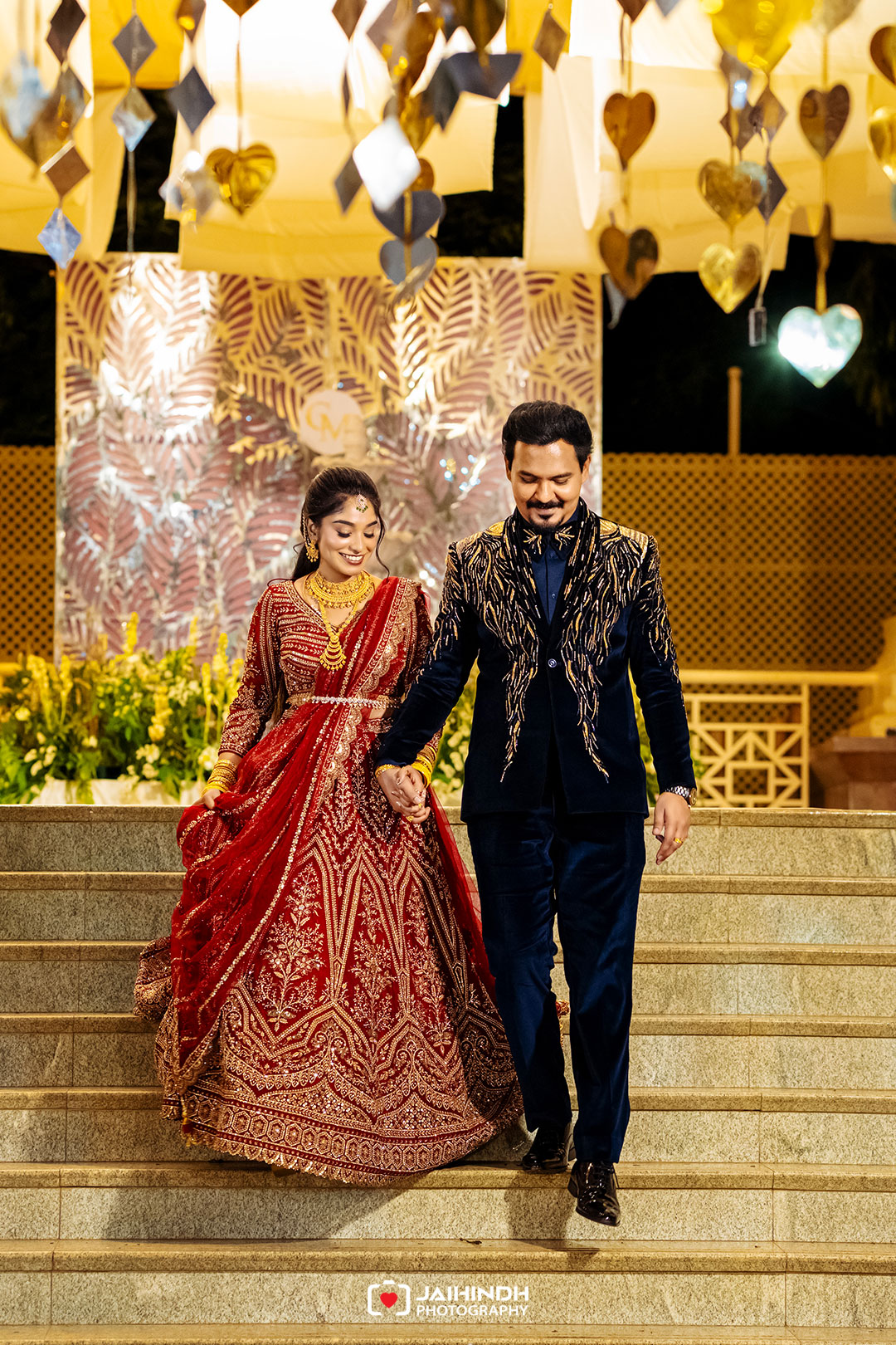 Bride in a red lehenga and groom in a navy sherwani walk down steps hand in hand at a festive decorated wedding venue.