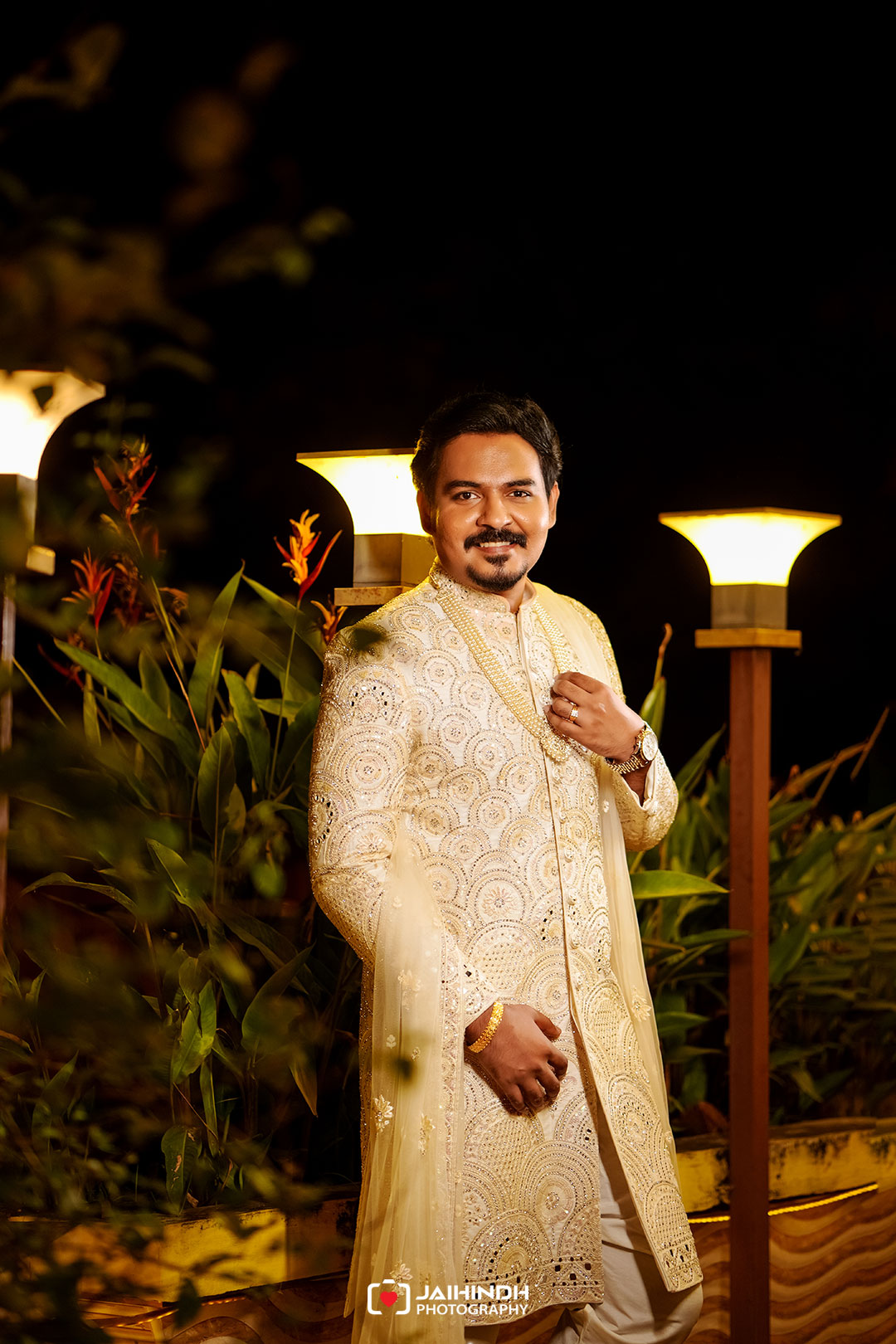 Man in an ornate cream sherwani stands outdoors at night, smiling, with warm lamp lights in the background.